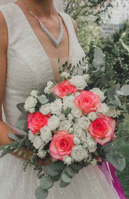 A bride holding a floral bouquet consisting of large, vibrant pink roses, smaller white roses, and gray-green eucalyptus leaves, arranged in a lush, rounded style. The bouquet is wrapped with purple r